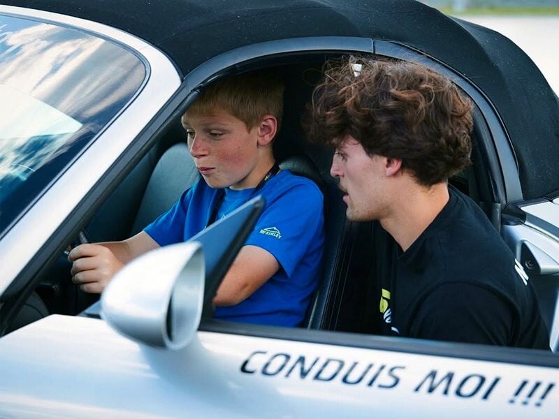 Un jeune garçon au volant d'une voiture blanche reçoit des conseils d'un instructeur assis à côté sur un circuit.