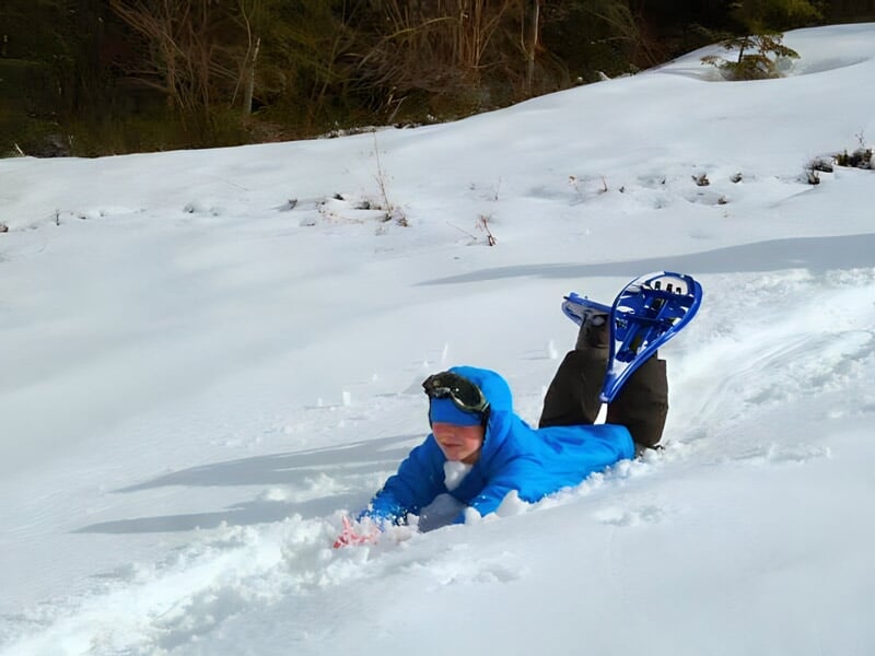 Enfant en combinaison bleue allongé dans la neige avec des raquettes aux pieds dans un paysage hivernal.