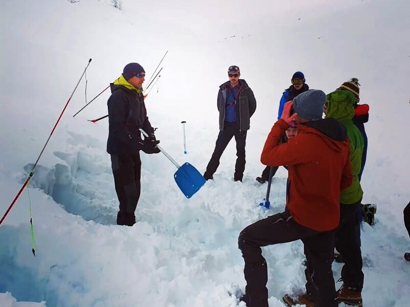 Groupe de personnes debout sur la neige avec pelles, discutant dans un cadre montagneux hivernal.