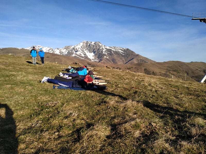 Personnes allongées sur un tapis dans une prairie avec montagnes en arrière-plan sous un ciel bleu.