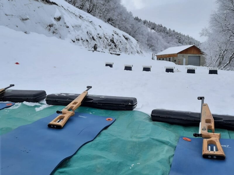 Carabines posées sur des tapis sur la neige devant un stand de tir avec un bâtiment en arrière-plan.