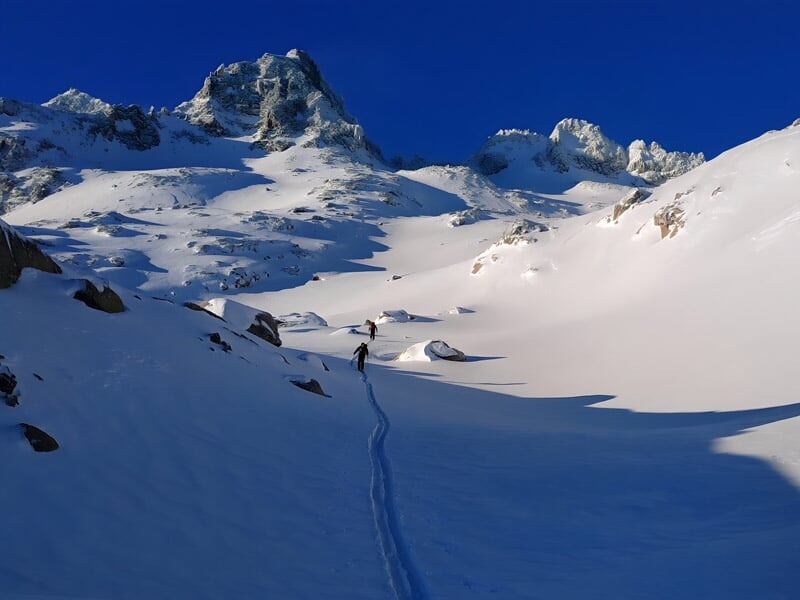 Deux personnes marchant dans une vallée enneigée avec montagnes en arrière-plan sous un ciel bleu.