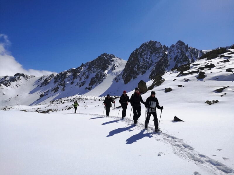 Groupe de cinq randonneurs progressant en file indienne sur neige immaculée, montagnes escarpées sous ciel bleu.
