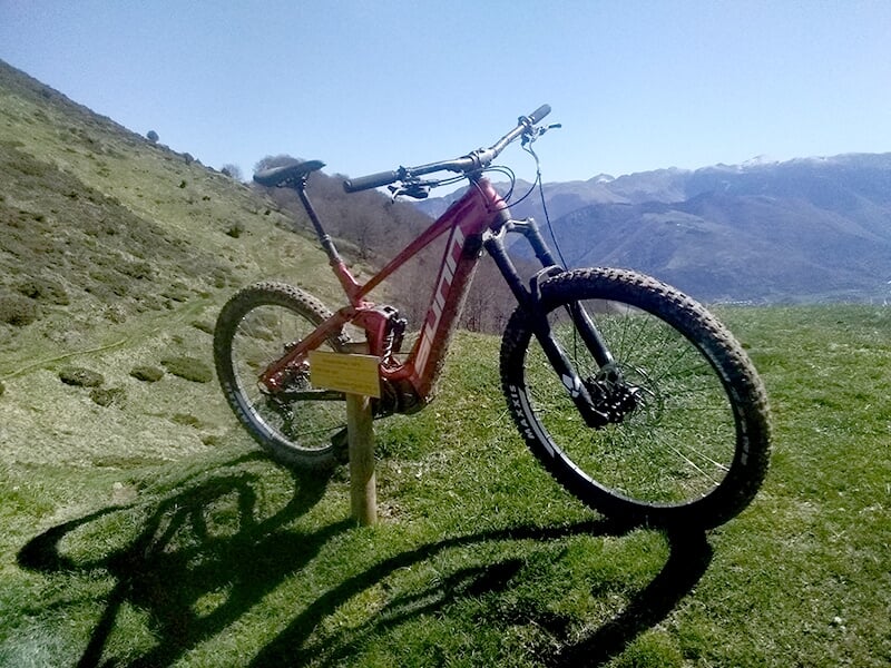 Un vélo tout-terrain rouge appuyé sur un panneau en bois, avec un paysage vallonné et un ciel dégagé en arrière-plan.