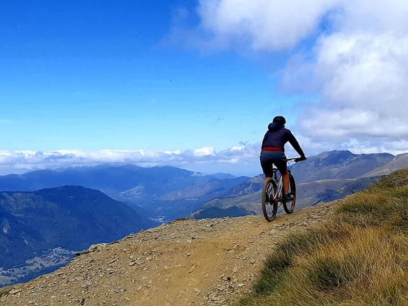 Un vététiste vêtu d'une tenue sombre pédale sur un sentier de montagne escarpé, avec une vue panoramique sur les sommets et les vallées environnantes.