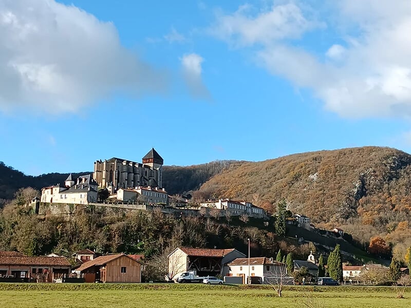 Vue d’un village médiéval avec un château perché sur une colline, entouré de montagnes et de verdure.