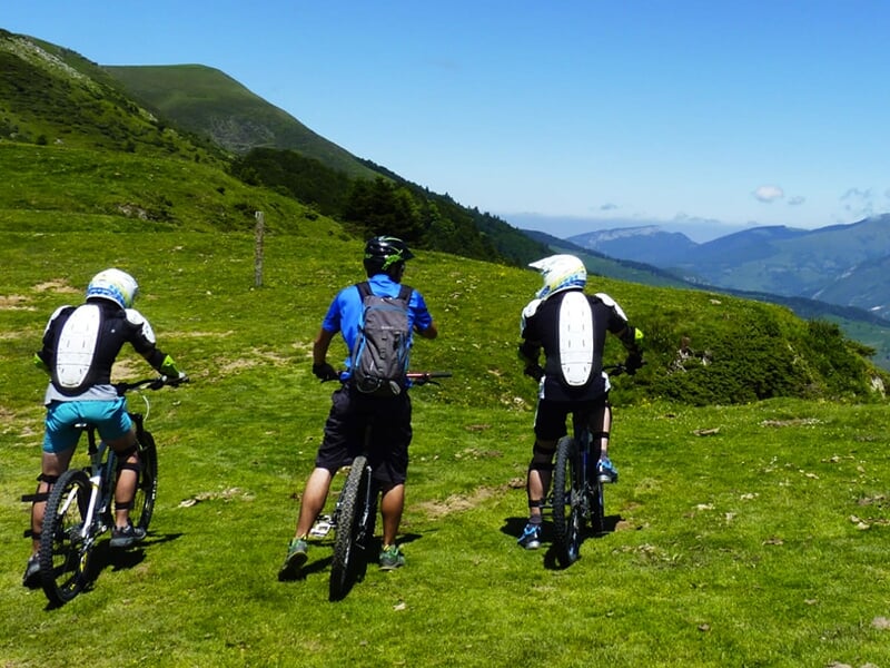 Trois cyclistes en tenue de protection se préparent à descendre un sentier de montagne verdoyant, encadrés par un guide portant un sac à dos.