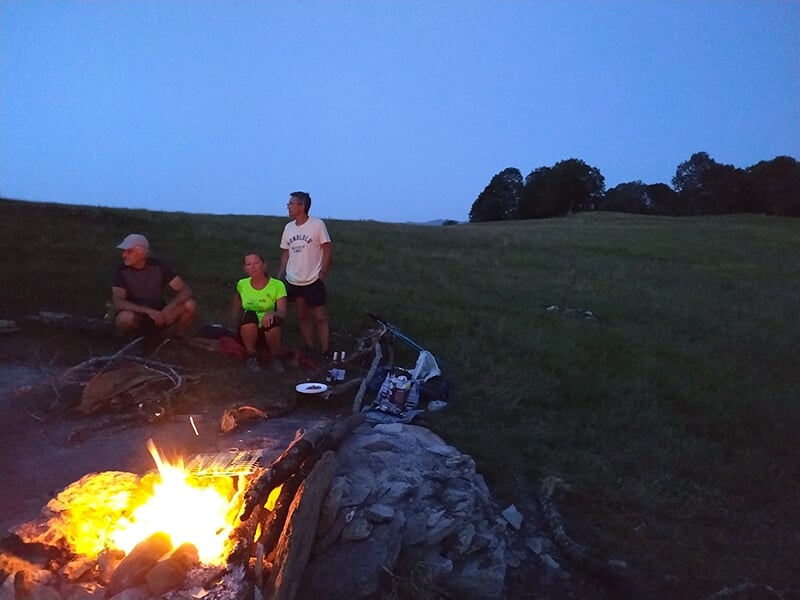 Trois personnes se détendent autour d’un feu de camp au crépuscule, sur une colline herbeuse, avec un ciel bleu nuit en arrière-plan.