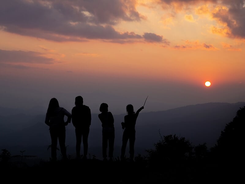 Silhouettes de quatre personnes sur une colline au coucher du soleil, avec des montagnes en arrière-plan et un ciel coloré.