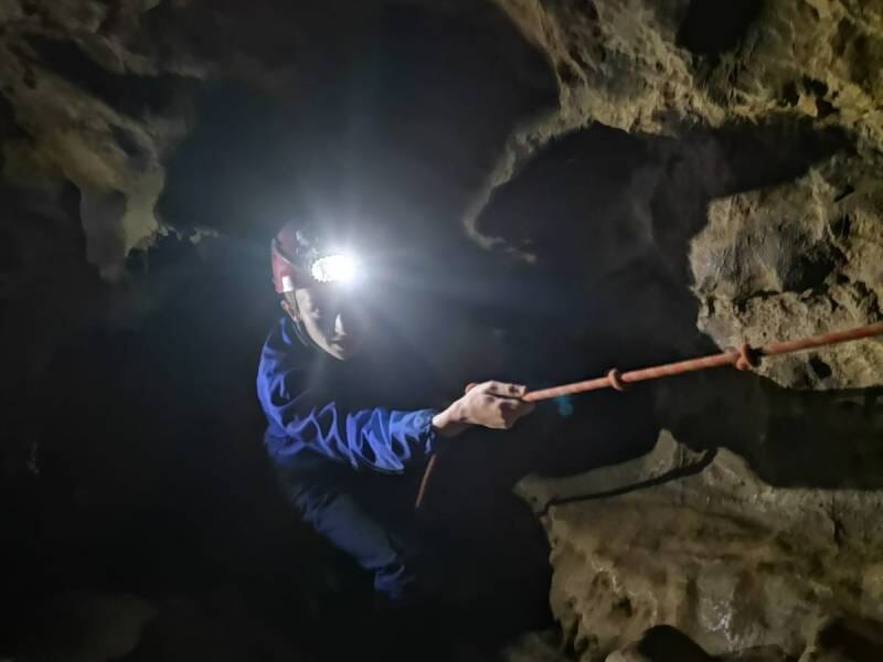 Une personne en combinaison bleue avec un casque et une lumière dessus, qui descend en rappel dans une grotte.