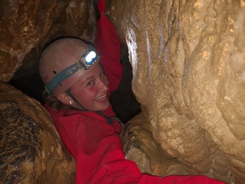 Une jeune fille en combinaison rouge et avec un casque qui se faufile dans un passage étroit.