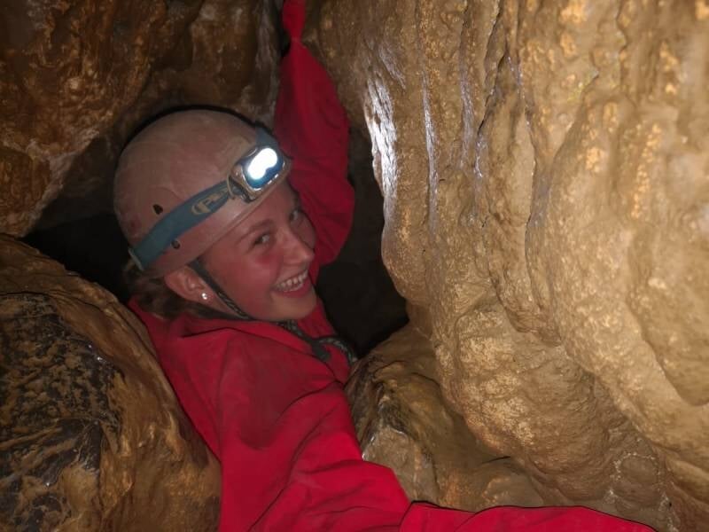 Une jeune fille en combinaison rouge et avec un casque qui se faufile dans un passage étroit.