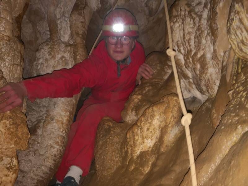 Un jeune homme vêtu d'une combinaison rouge et d'un casque avec une lampe qui avance dans une grotte étroite.