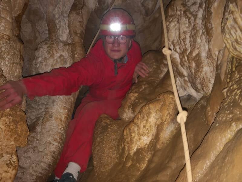 Un jeune homme vêtu d'une combinaison rouge et d'un casque avec une lampe qui avance dans une grotte étroite.