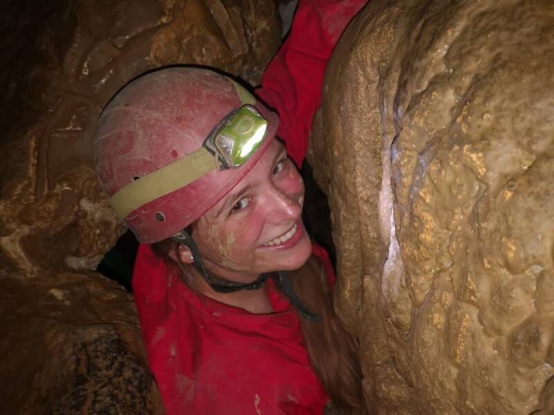 Une jeune femme souriante, vêtue d'une combinaison et d'un casque rouges, dans un passage étroit.