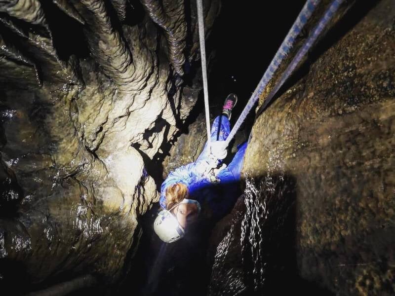 Une jeune personne vêtue d'une combinaison bleue qui descend en rappel dans une grotte.
