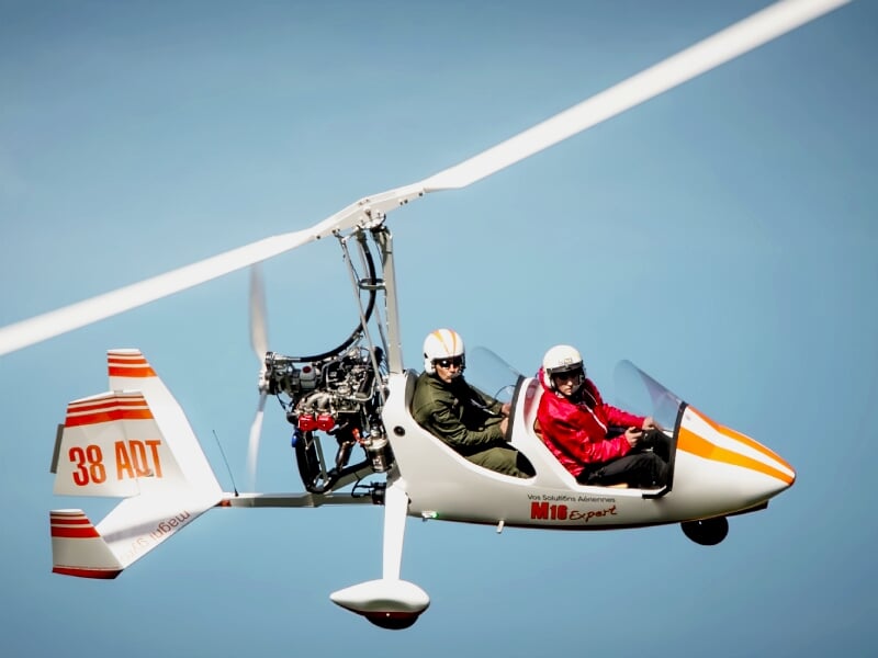 un ULM autogire orange et blanc en plein vol avec deux passagers