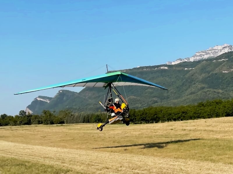 Personne en vol en ULM pendulaire au-dessus d'un champ avec montagnes en arrière-plan sous un ciel bleu clair.