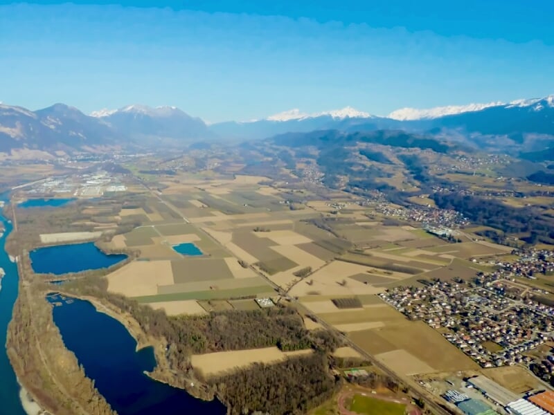 Vue aérienne d'une vallée avec champs cultivés, plans d'eau et montagnes enneigées à l'horizon sous un ciel clair.
