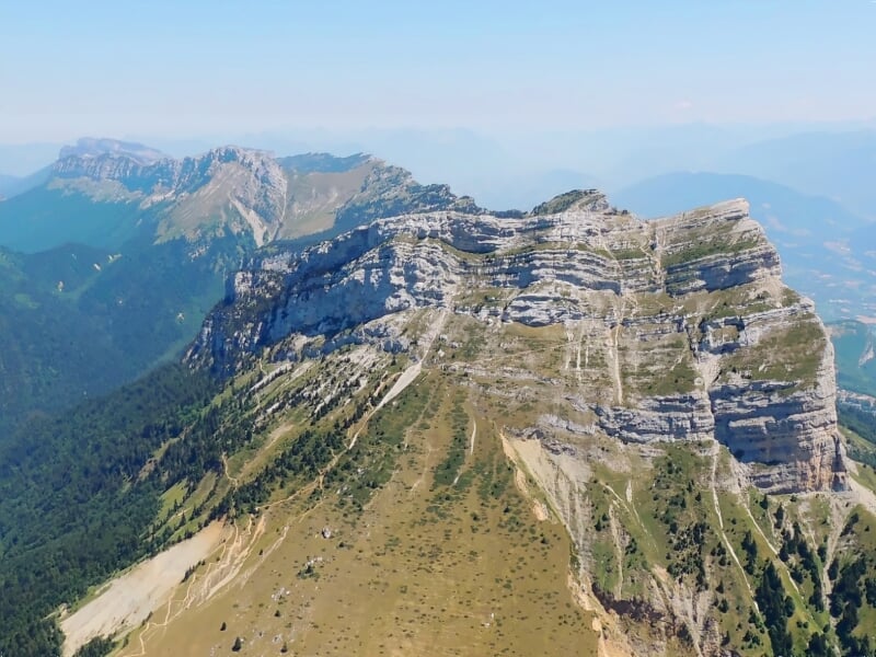 Montagne rocheuse imposante avec des strates visibles, entourée de végétation verte sous un ciel bleu clair.