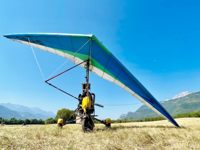 ULM pendulaire posé dans un champ sec avec montagnes en arrière-plan sous un ciel bleu lumineux et dégagé.