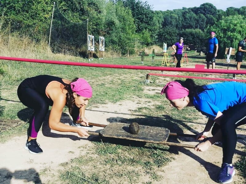 Deux femmes en tenue de sport soulèvent une planche avec un objet lourd dessus dans un parc ensoleillé.