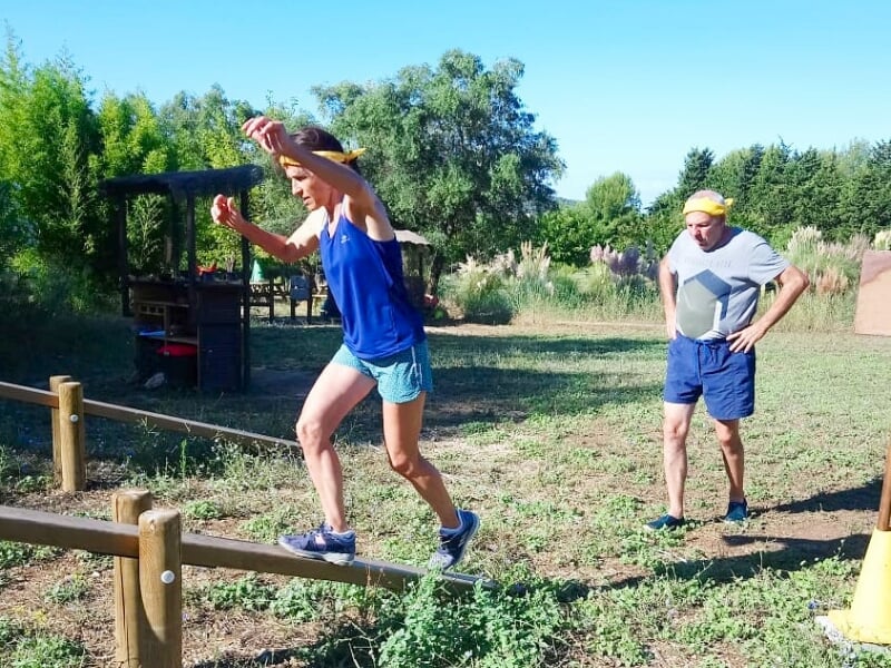 Femme en short bleu marche sur une poutre en bois pendant qu'un homme l'observe dans un espace naturel.