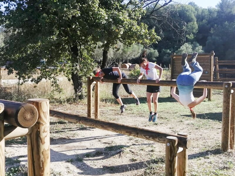 Trois personnes franchissent une barre horizontale en bois dans un parcours d'obstacles en extérieur.