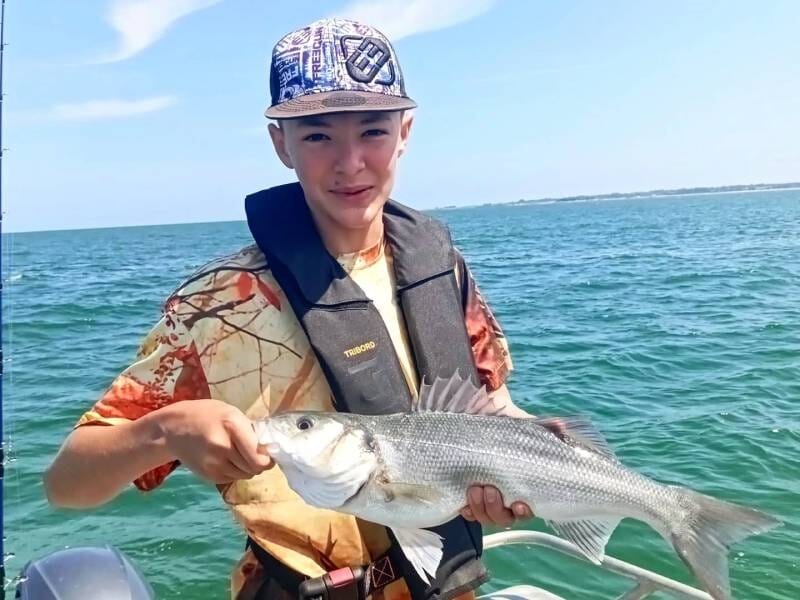 Un jeune garçon en gilet de sauvetage tient un poisson blanc sur un bateau en mer par une journée ensoleillée.