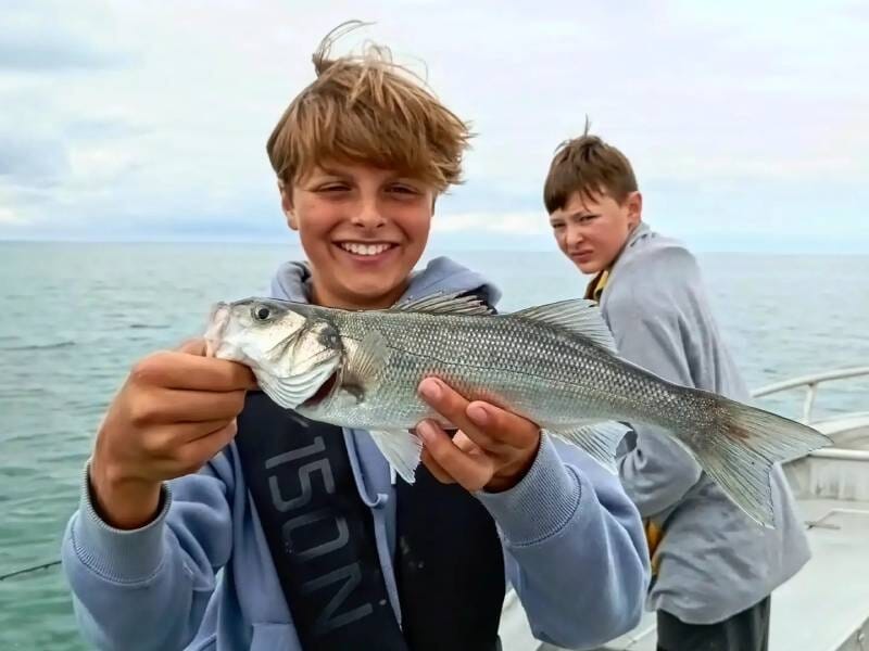 Deux garçons sur un bateau en mer, l'un souriant montre un poisson blanc qu'il vient de pêcher, près de Grandcamp-Maisy.