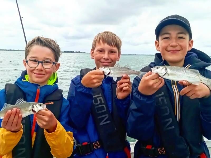 Trois garçons en gilets de sauvetage tiennent chacun un petit poisson blanc sur un bateau en mer sous un ciel nuageux.