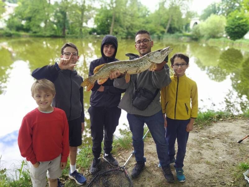 Un groupe de cinq personnes, dont un moniteur, posent au bord d'un étang avec un grand brochet attrapé lors d'un stage de pêche, près de Viry-Châtillon.