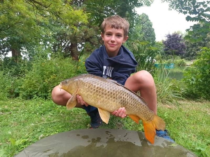Un garçon souriant tient une grosse carpe dorée assis sur l'herbe verte lors d'un stage de pêche en plein air, près de Viry-Châtillon.
