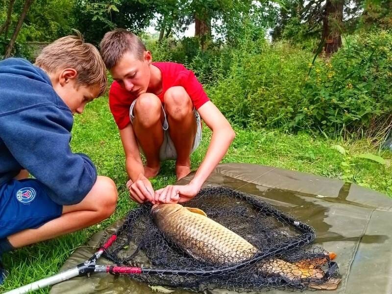 Deux garçons examinent une grosse carpe posée sur un tapis de pêche, entourés de végétation dense et d'arbres, près de Viry-Châtillon.