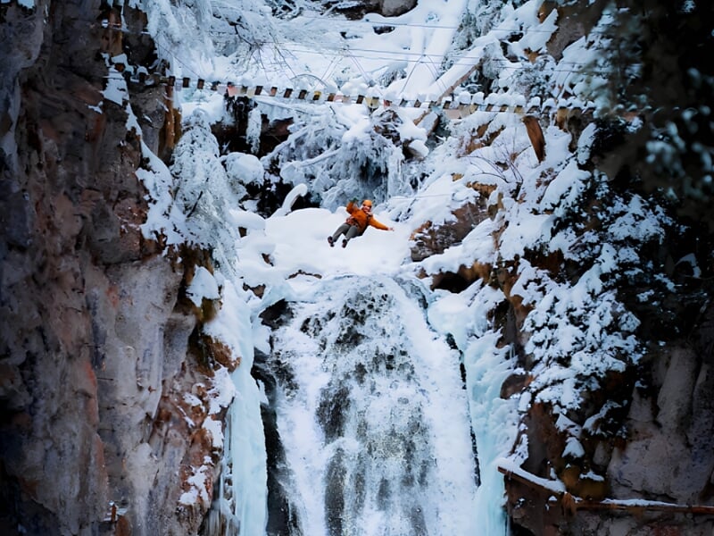 Personne en tenue orange sautant au-dessus d'une cascade gelée dans un paysage montagneux enneigé à Bagnères-de-Bigorre.