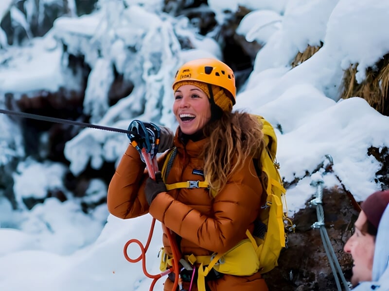Femme souriante en casque jaune et veste orange tenant une corde d'escalade dans un décor hivernal enneigé.