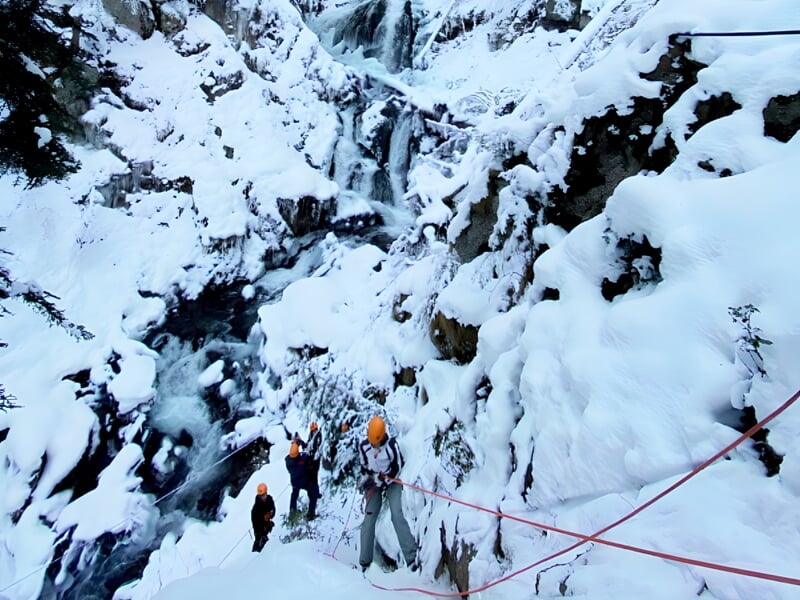 Groupe de personnes équipées pour via ferrata progressant sur un sentier enneigé au bord d'une rivière glacée.