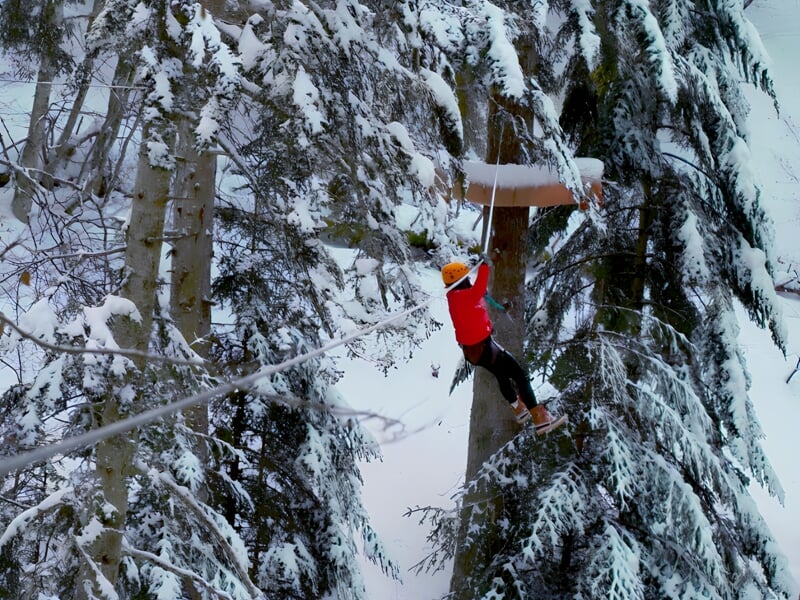 Personne en veste rouge et casque jaune traversant une tyrolienne au milieu d'une forêt enneigée à Bagnères-de-Bigorre.