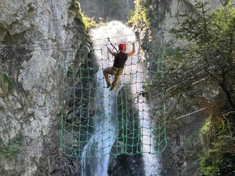 Un homme grimpe un filet devant une cascade pour traverser un cours d'eau en via ferrata