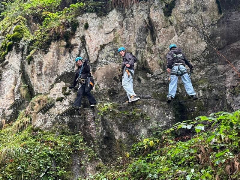 Trois personnes faisant de la via ferrata à flanc de falaise. Pierre, feuille et mousse se mélangent