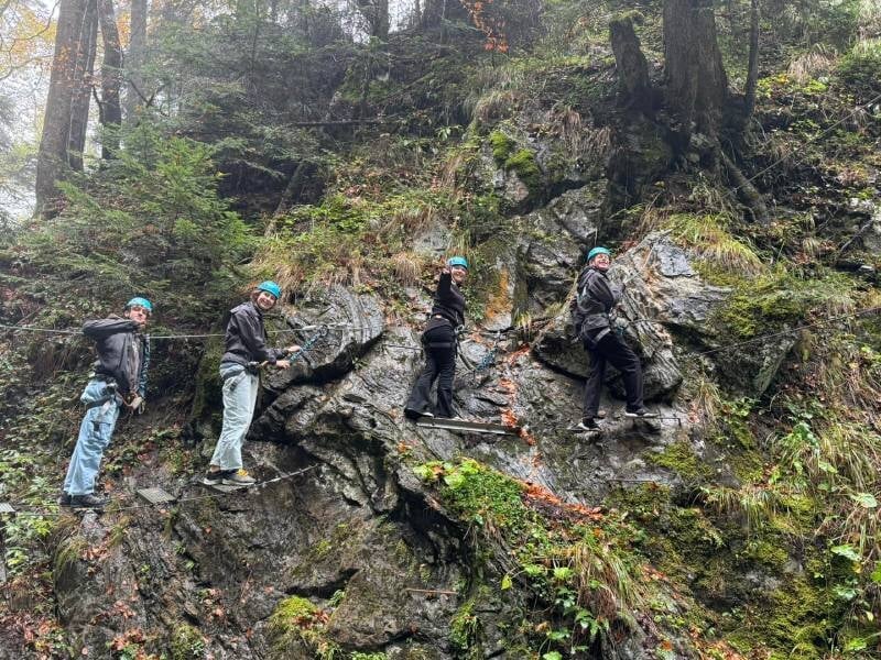 Quatre personnes souriantes faisant de la via ferrata à flanc de falaise. Pierre, feuille et mousse se mélangent