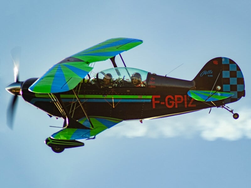 Avion de voltige biplan vert et bleu en vol avec deux personnes à bord sous un ciel clair