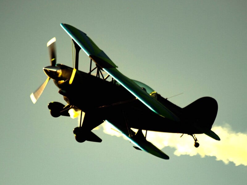 Avion biplan noir en vol avec une traînée de fumée blanche sur fond de ciel clair au coucher du soleil