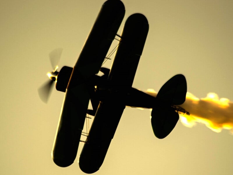 Avion biplan noir en vol avec une traînée de fumée orange sur fond de ciel au coucher du soleil