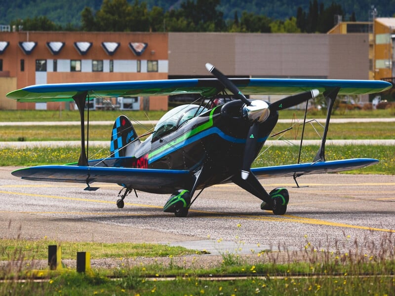 Avion de voltige biplan bleu et vert stationné sur une piste d'aéroport avec des bâtiments en arrière-plan