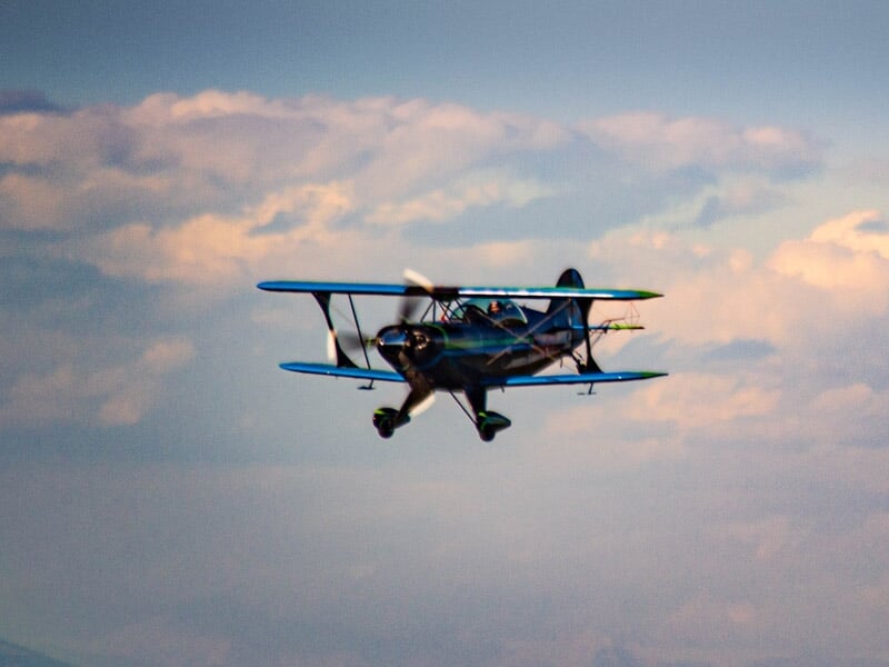 Avion biplan bleu et vert en vol face à l'appareil avec un ciel nuageux en arrière-plan