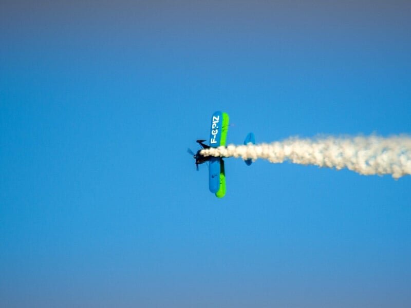 Avion de voltige bleu et vert en vol vertical avec une traînée de fumée blanche sur fond de ciel bleu