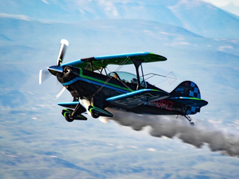 Avion biplan bleu et vert en vol avec deux personnes à bord et une traînée de fumée noire derrière