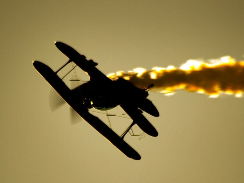 Avion biplan noir en vol avec une traînée de fumée orange sur fond de ciel au coucher du soleil