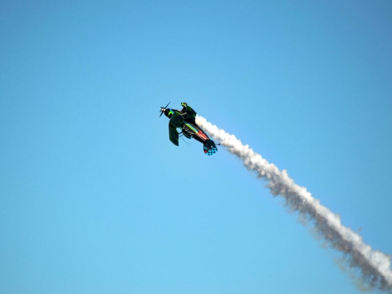 Avion de voltige vert et noir en vol vertical avec une traînée de fumée blanche sur fond de ciel bleu